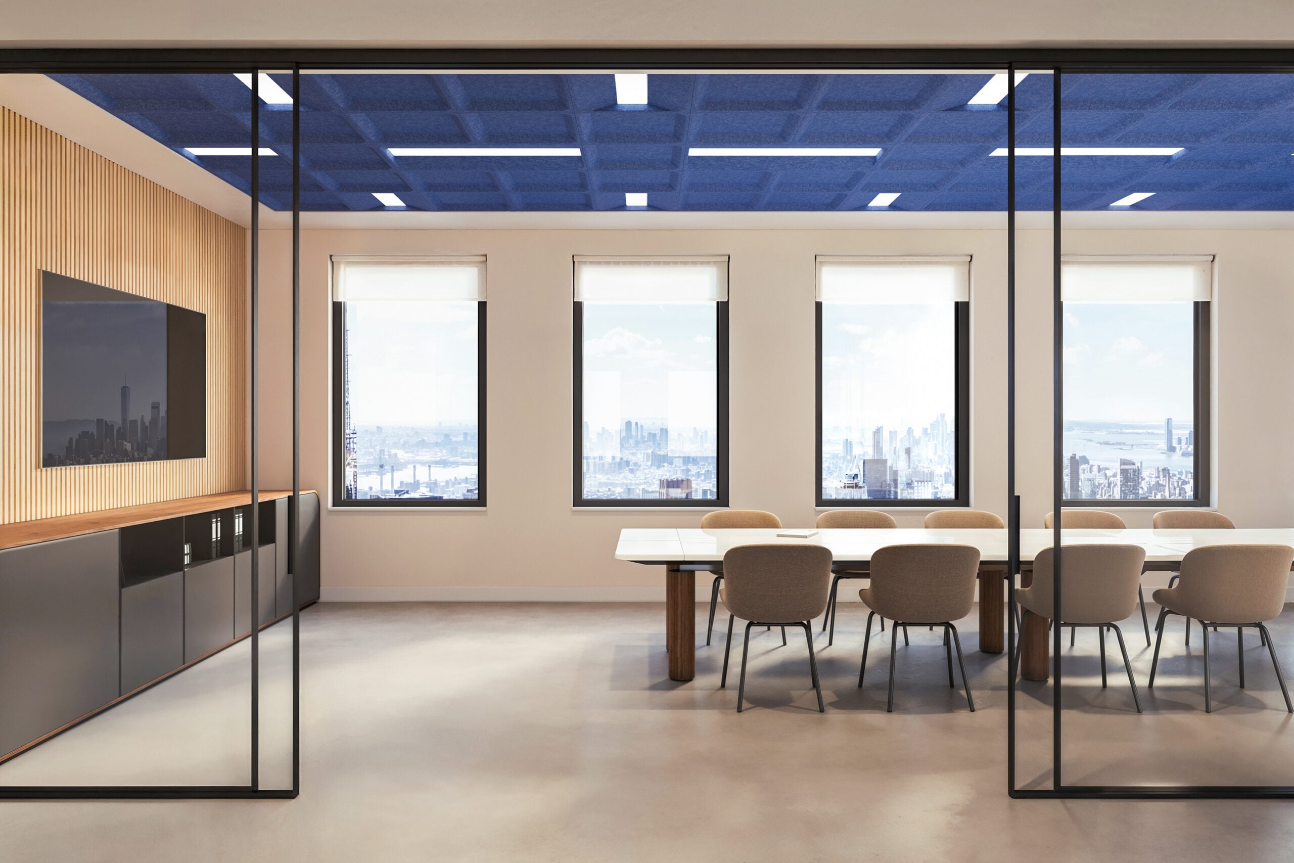Modern conference room with large windows, a long table surrounded by chairs, a wall-mounted screen, and city skyline visible outside.