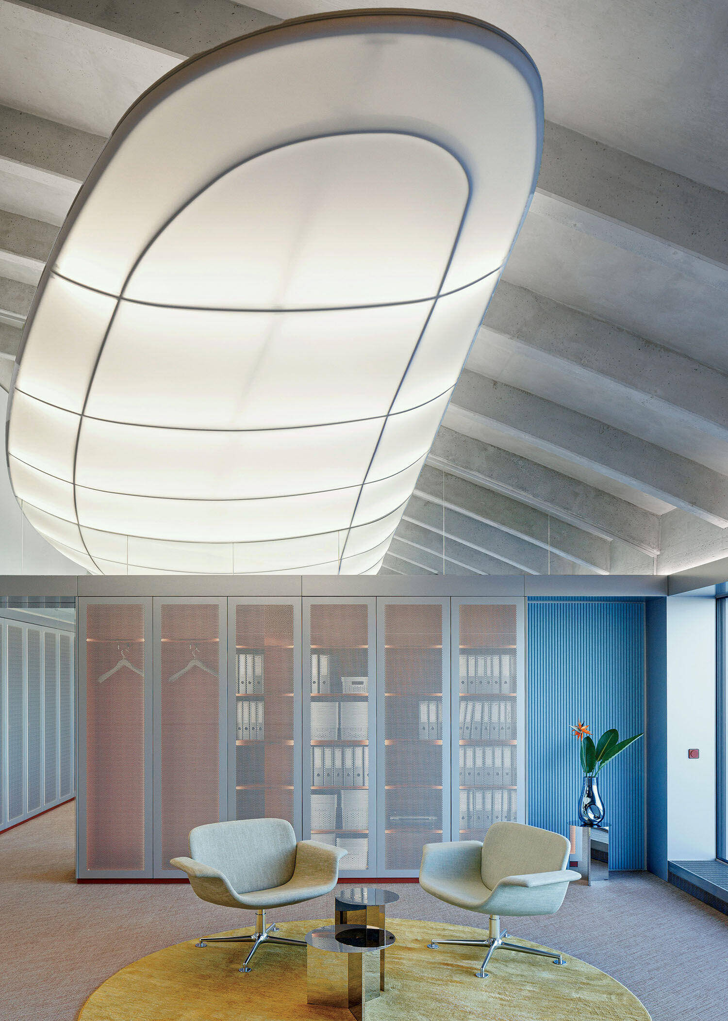 Modern office lounge with two beige chairs, a small round glass table, built-in shelving with documents, and a large, curved ceiling light fixture.