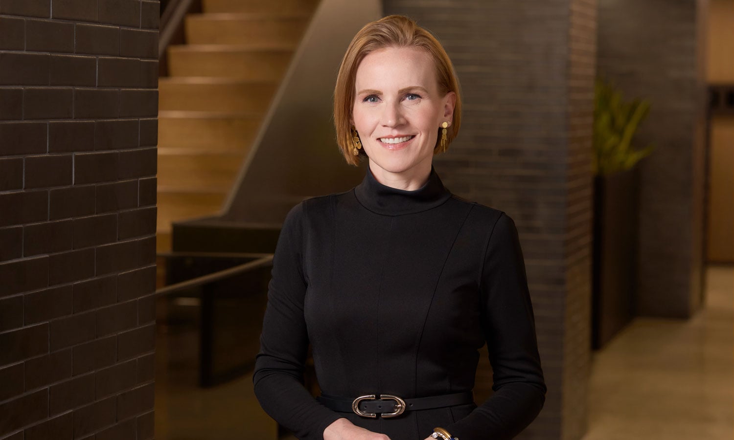 A woman with short blonde hair, wearing a black long-sleeve dress, stands indoors in front of a staircase and brick wall, smiling at the camera.