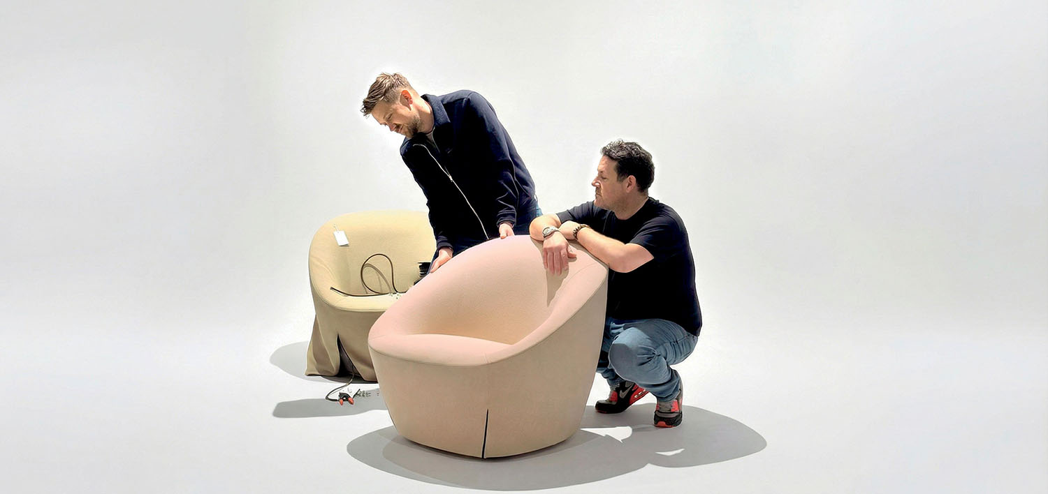 Two men examine and adjust a light pink armchair on a white background; a beige armchair with visible cords sits nearby.