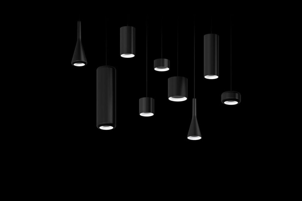 Nine modern black pendant lights of varying lengths and shapes hang against a solid black background, with the bulbs facing downward.