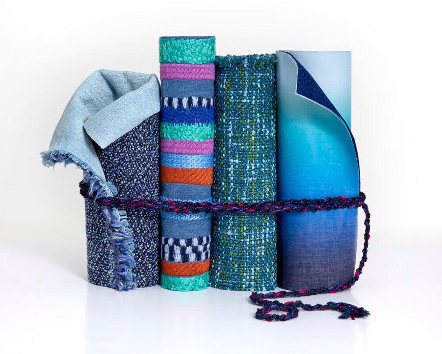 Four rolls of assorted textured and patterned fabric, standing upright and tied together with a braided cord against a white background.