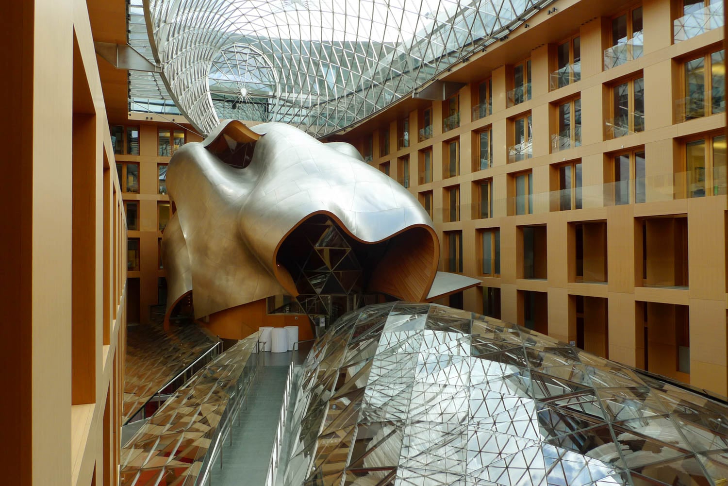 A modern atrium with a large, abstract metallic sculpture and geometric glass ceilings, surrounded by wood-paneled walls and windows.