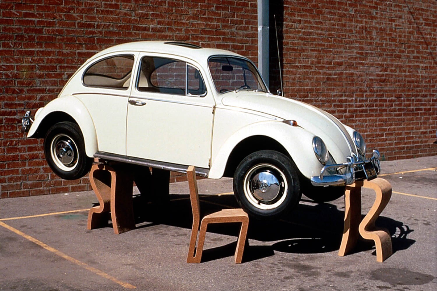 A white Volkswagen Beetle is elevated off the ground, supported by several wooden chairs in a parking lot next to a brick wall.