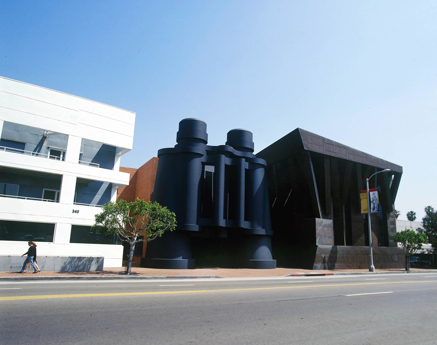 A large building with a giant pair of black binoculars at its entrance, flanked by modern white and dark angular structures, with a person walking on the sidewalk.