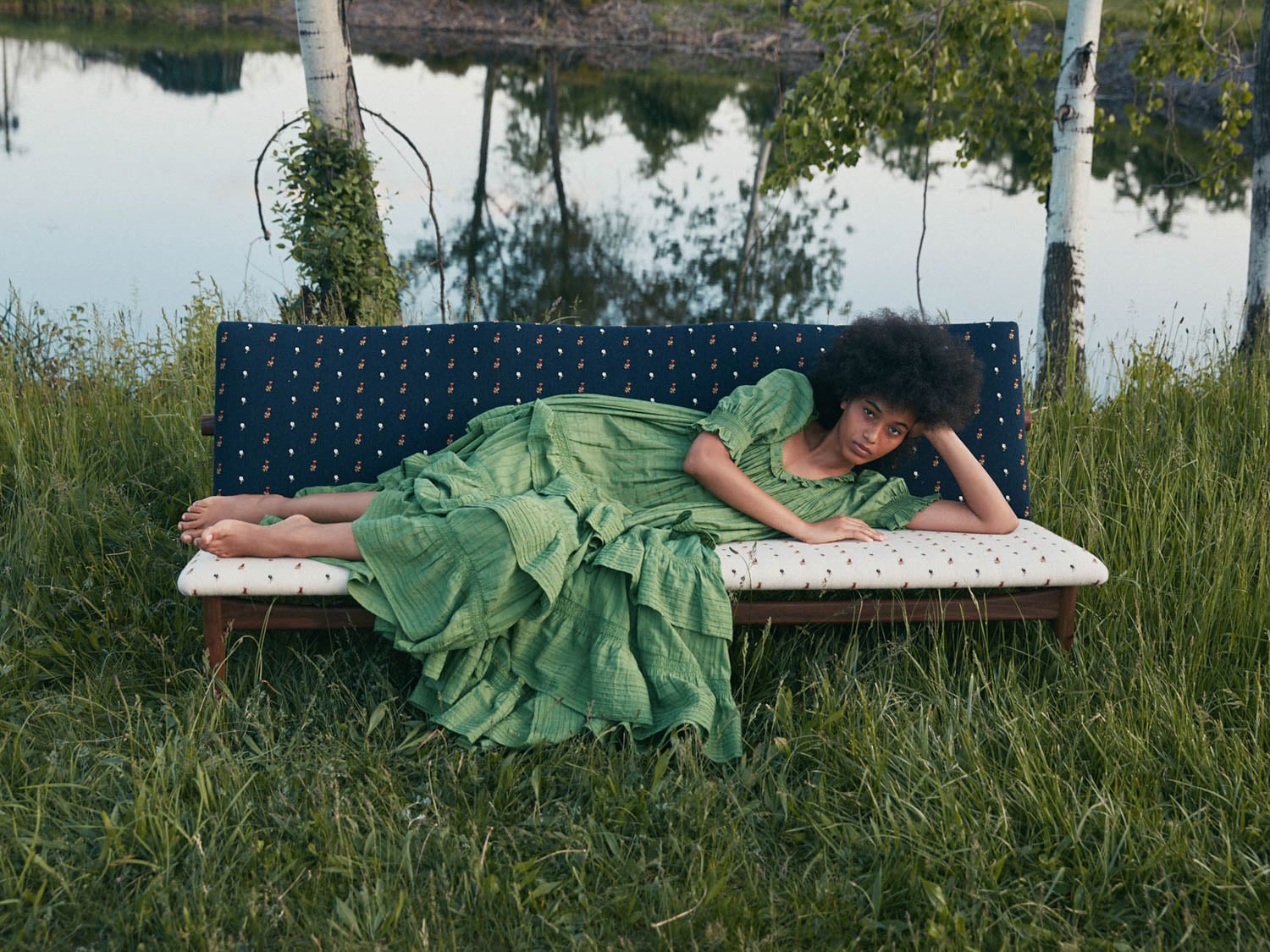 A person in a green dress lies on a cushioned bench outdoors, surrounded by grass and trees near a pond.