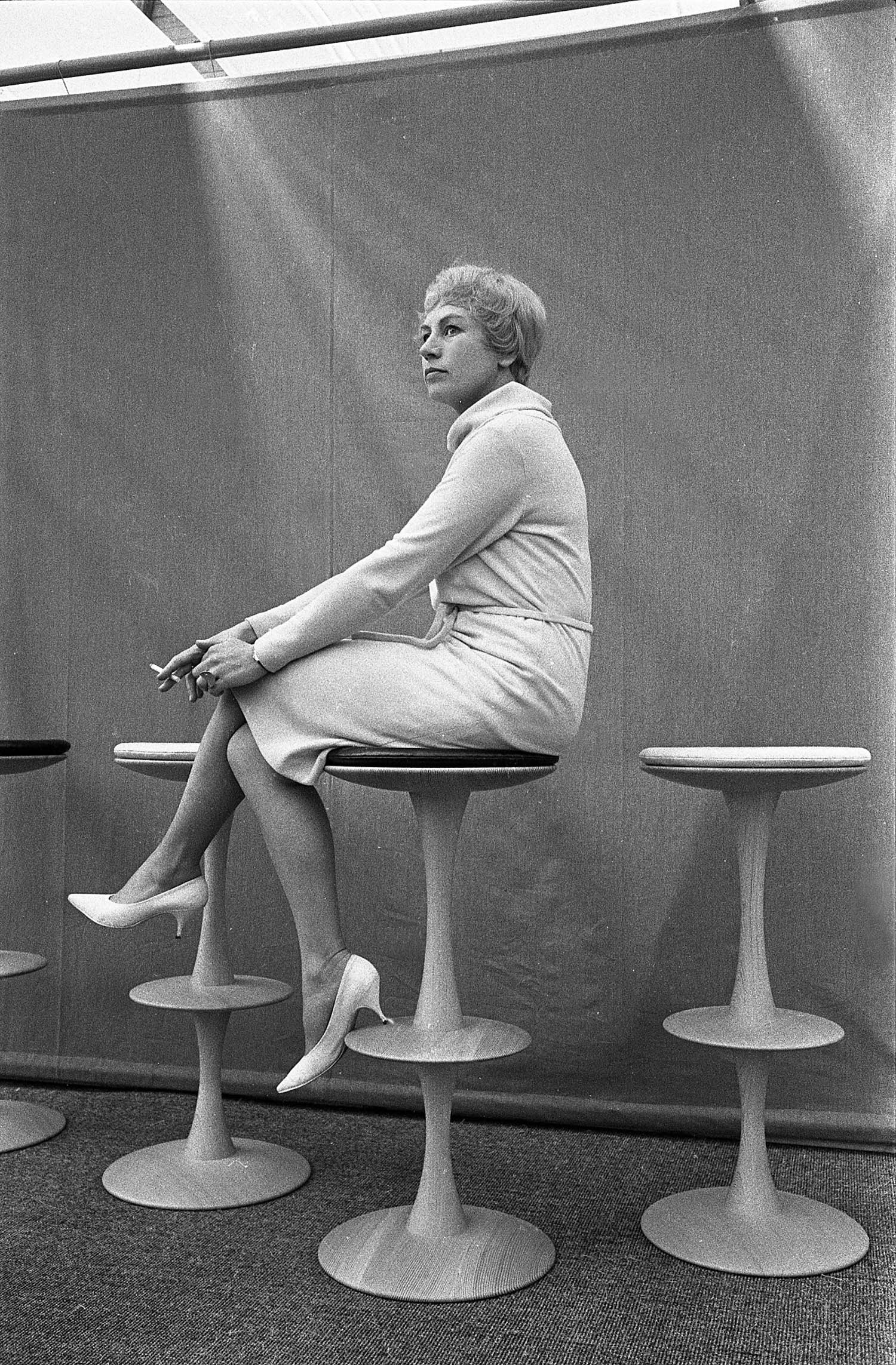 A woman in a light-colored outfit sits sideways on a high stool, holding a cigarette, with two empty stools beside her against a plain backdrop.