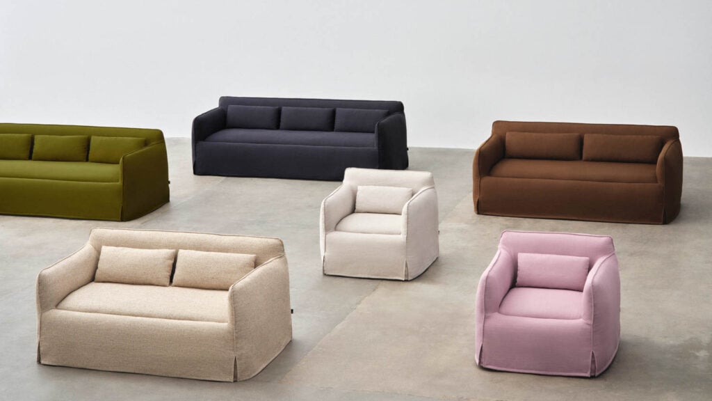 A group of modern sofas and armchairs in various colors, including green, navy, brown, beige, and pink, arranged on a concrete floor against a plain white background.