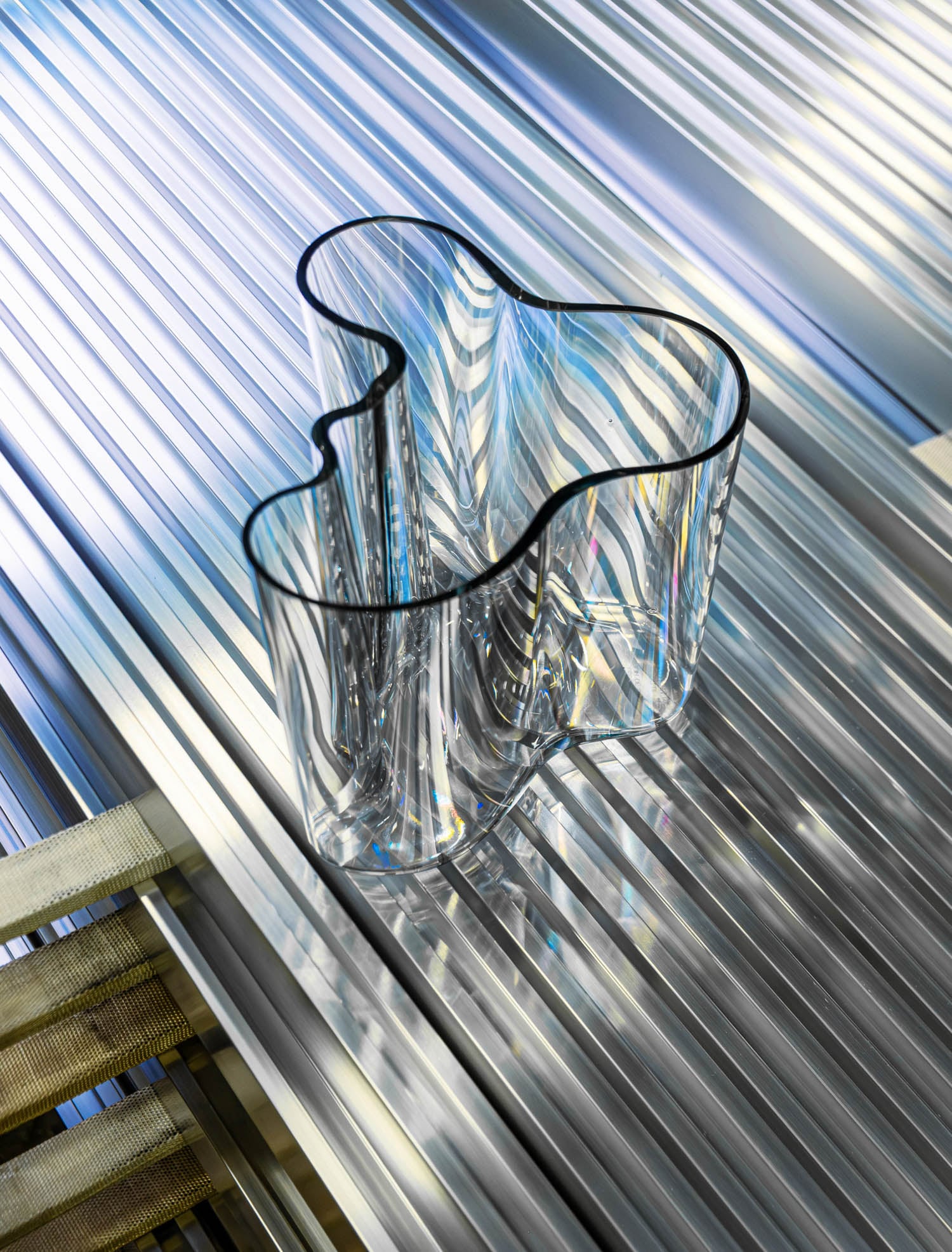 A clear, wavy-edged glass vase sits on a reflective, ribbed metallic surface with light creating colorful reflections and shadows.