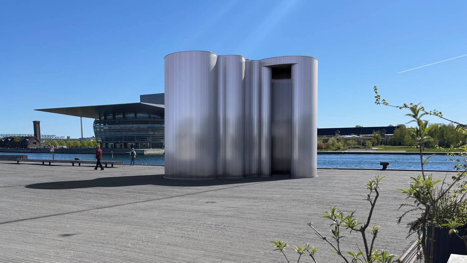 A modern, cylindrical metallic structure stands on a waterfront promenade with a large glass building and water in the background under a clear blue sky.