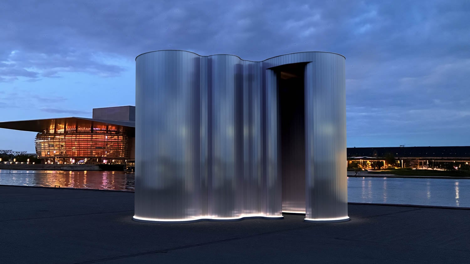 A wavy, metallic pavilion with illuminated base stands near a waterfront at dusk; a large, lit building is visible across the water.