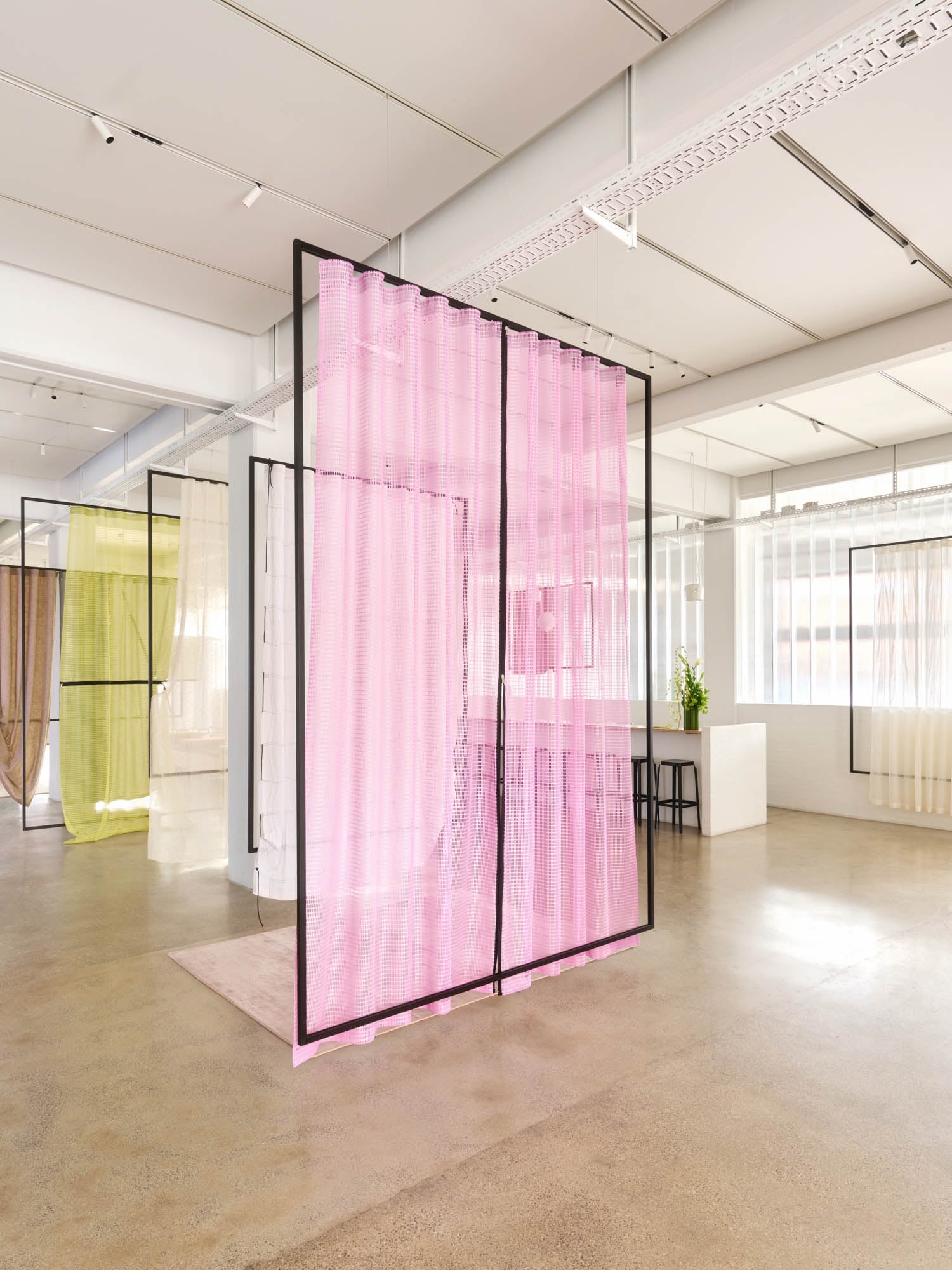 A modern interior with translucent colored fabric panels in black frames, featuring pink, yellow, and beige sections, on a polished concrete floor with ample natural light.