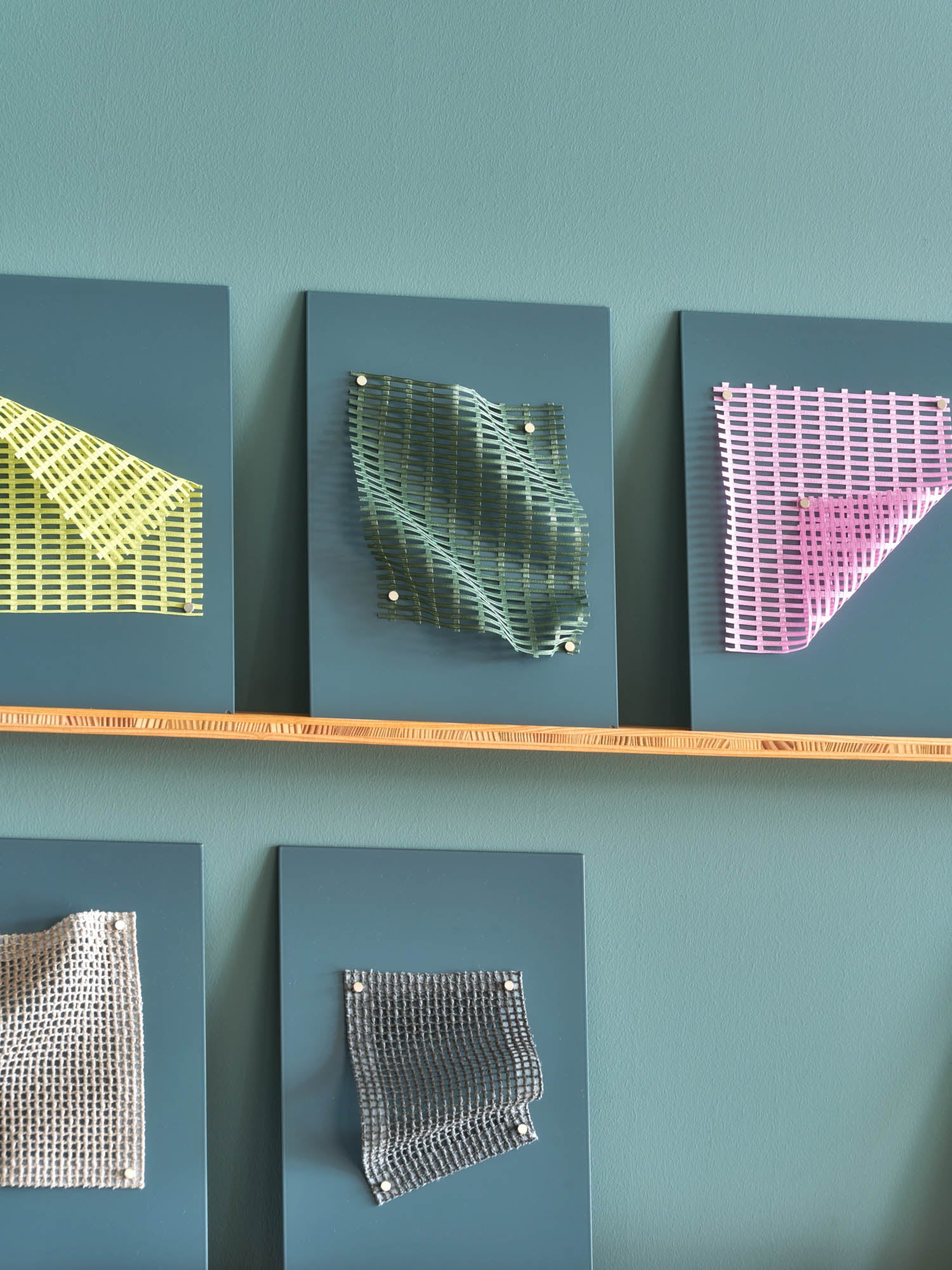 Five pieces of textured and patterned fabric are mounted on blue rectangular boards, displayed on a wooden shelf against a teal wall.