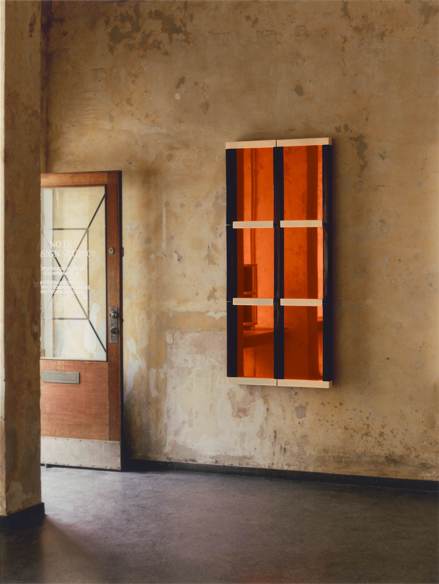 A wooden door and a wall-mounted cabinet with orange glass panels are set against a textured, beige wall in a minimalist interior.