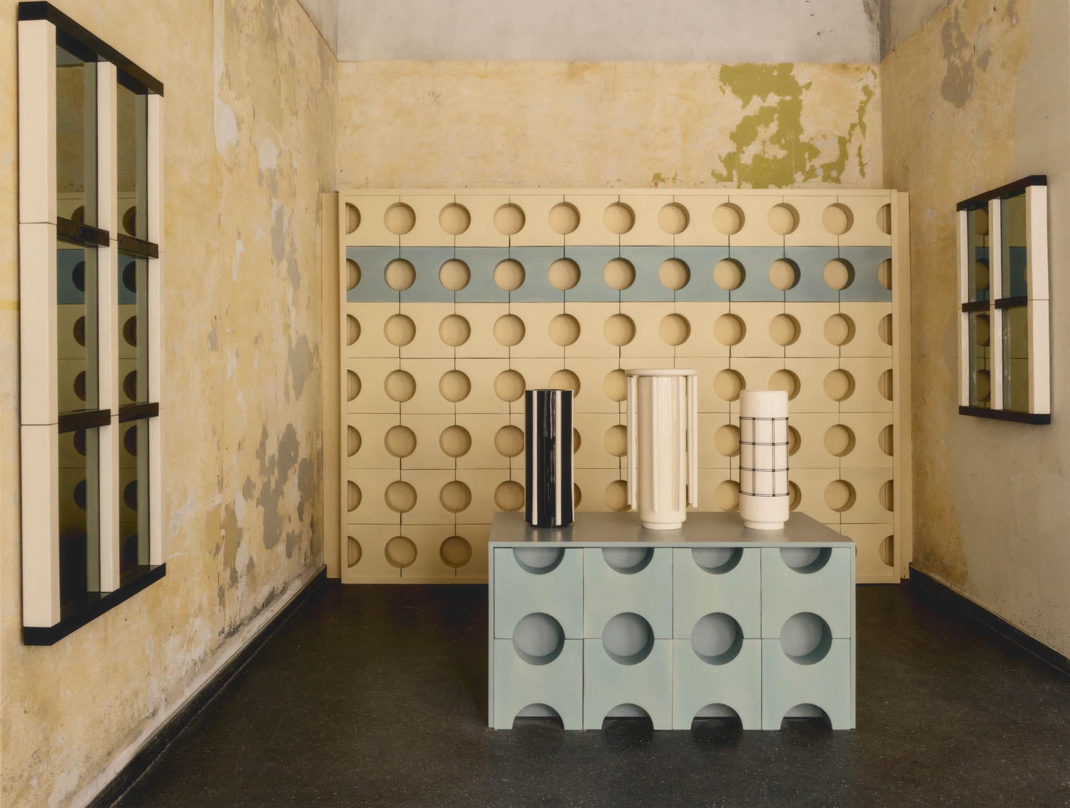 A room with worn yellow walls features geometric furniture, including a grid-patterned shelving unit, a blue table, three column-like vases, and two rectangular mirrors.