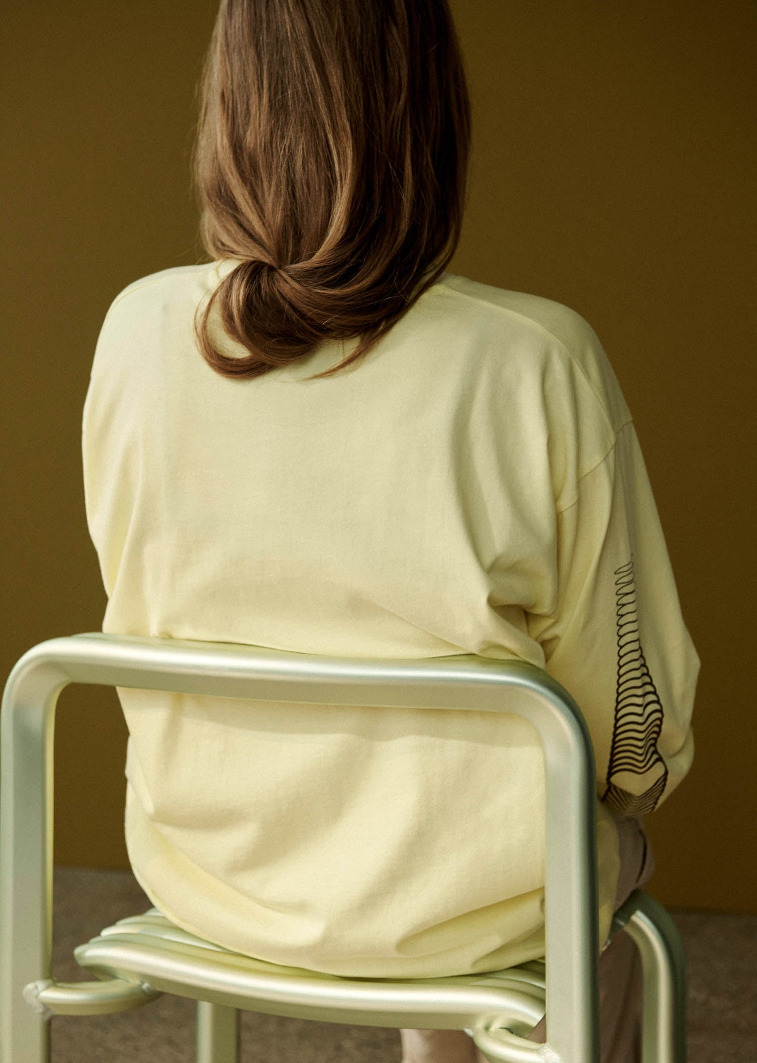 Person with long brown hair sits on a pale green chair, facing away from the camera, wearing a light yellow shirt with a black line design on the sleeve.
