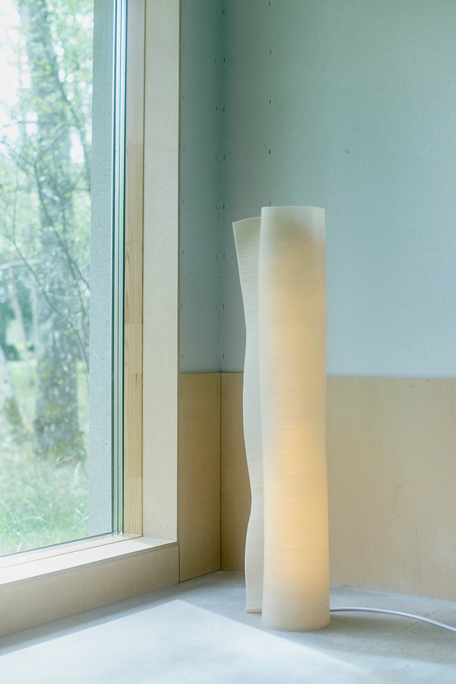 A tall, sculptural floor lamp with a soft white glow stands next to a large window in a minimalist room with pale walls and light wood paneling.