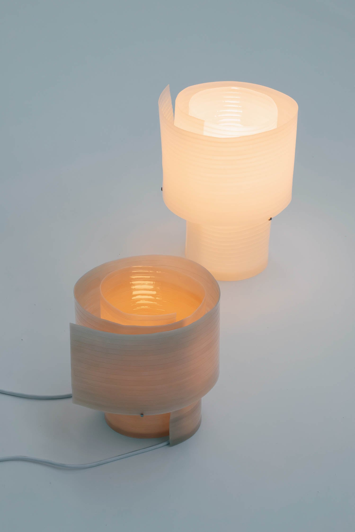 Two modern table lamps with cylindrical, layered translucent shades emitting warm, diffused light, placed on a light surface with one lamp plugged in.