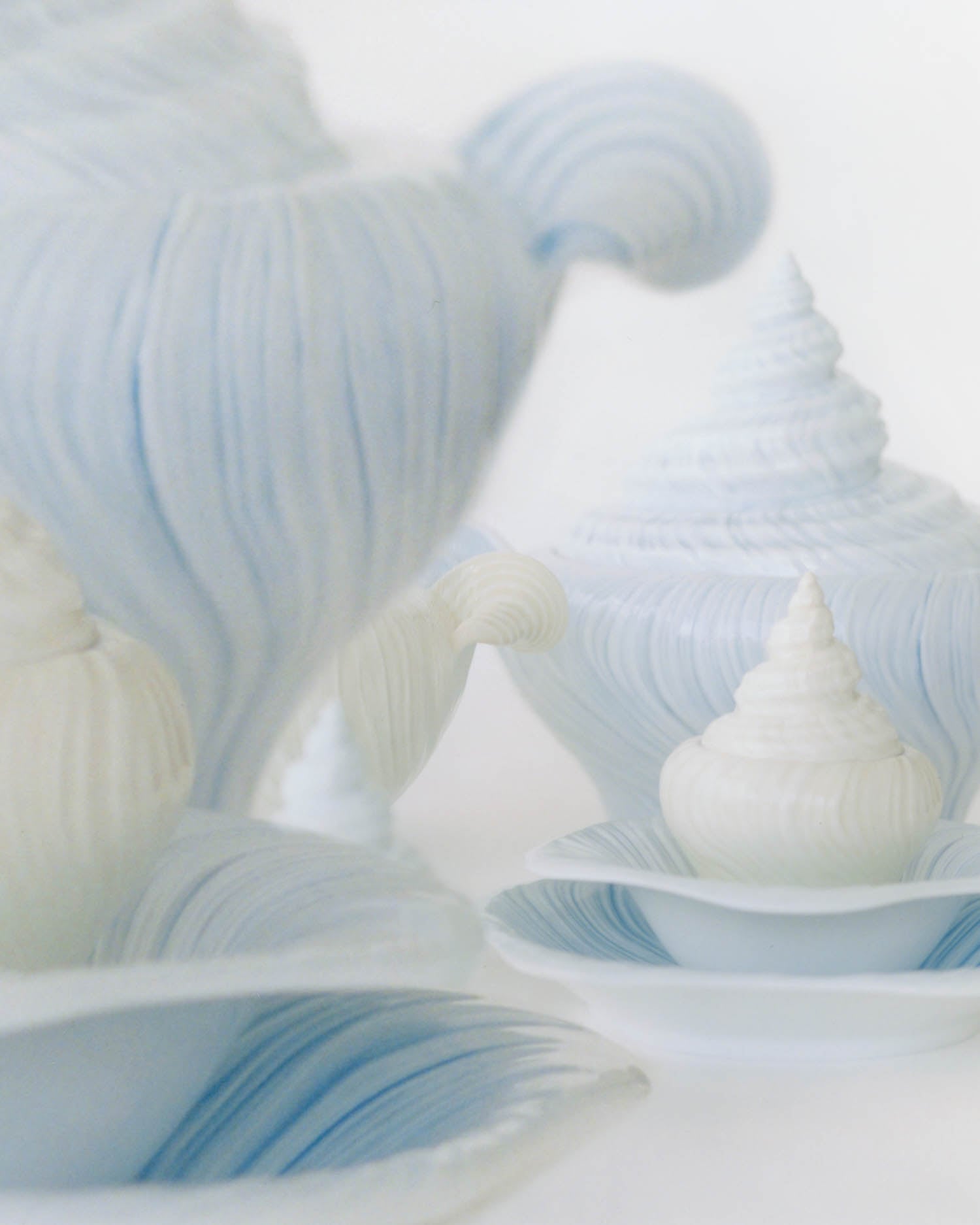 Close-up of intricately sculpted teacups and teapots with shell-like, wavy textures in pale blue and white on matching saucers against a white background.