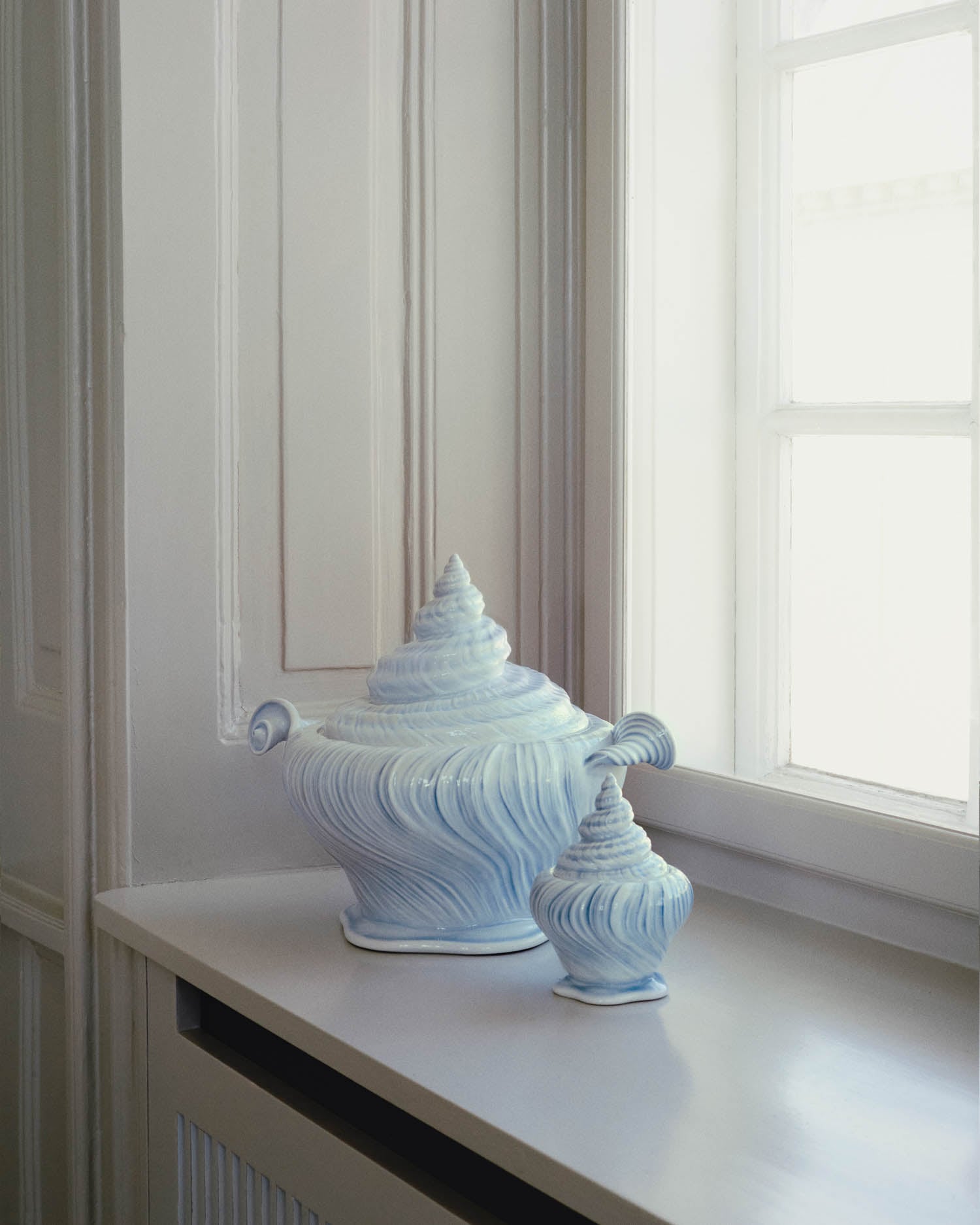 Two ceramic vases shaped like seashells, one large and one small, rest on a white windowsill beside a window with natural light.