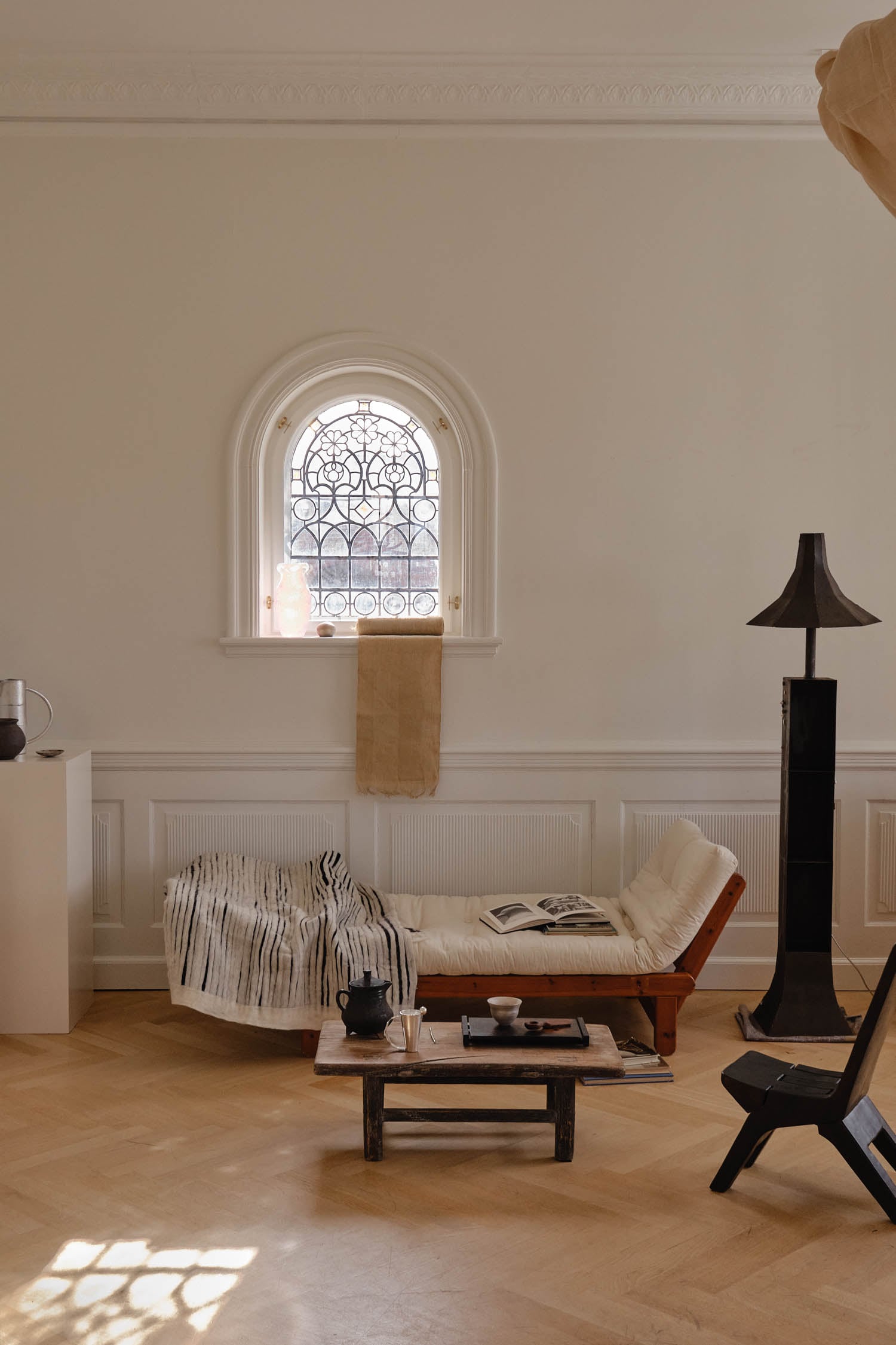 Minimalist living room with a daybed, striped throw, and low coffee table. An arched window with ornate glass lets in light. Neutral tones and simple decor create a calm atmosphere.