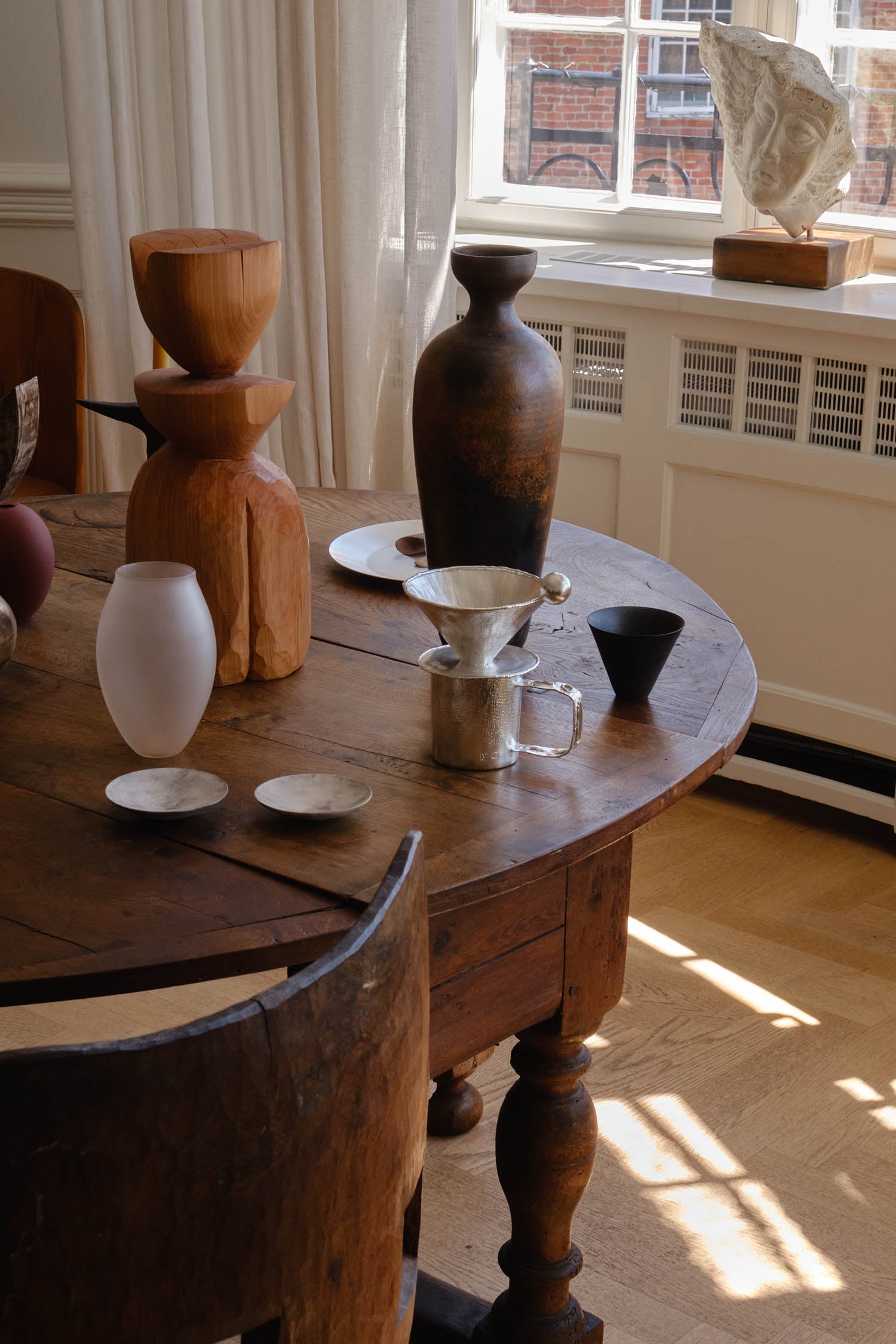 A wooden table with carved wooden bowls, ceramic vases, a metal pour-over coffee maker, and small plates near a sunlit window.