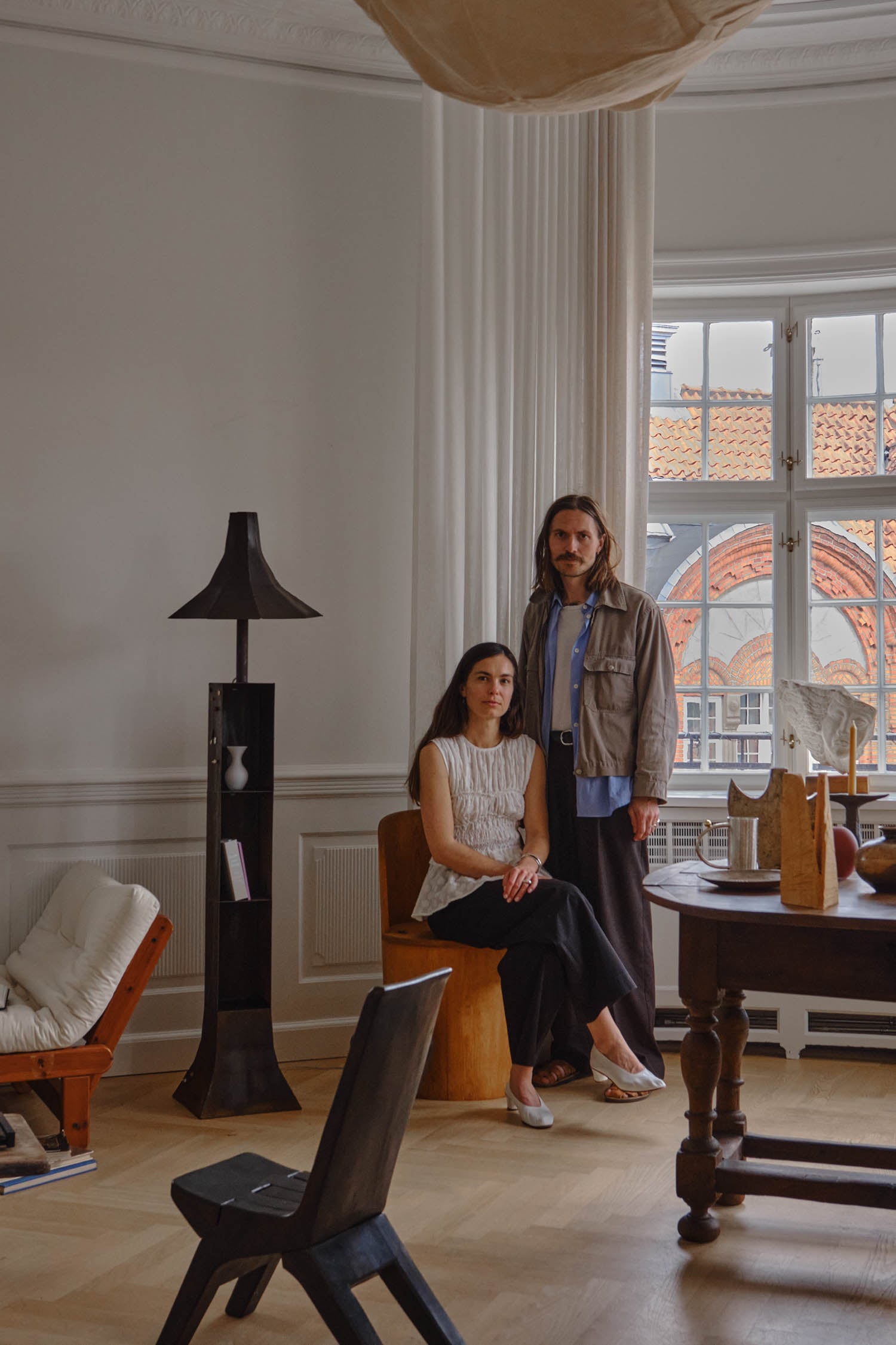 Two people pose in a modern, minimalist living room with wooden furniture, white walls, large windows, and decorative objects on the table.
