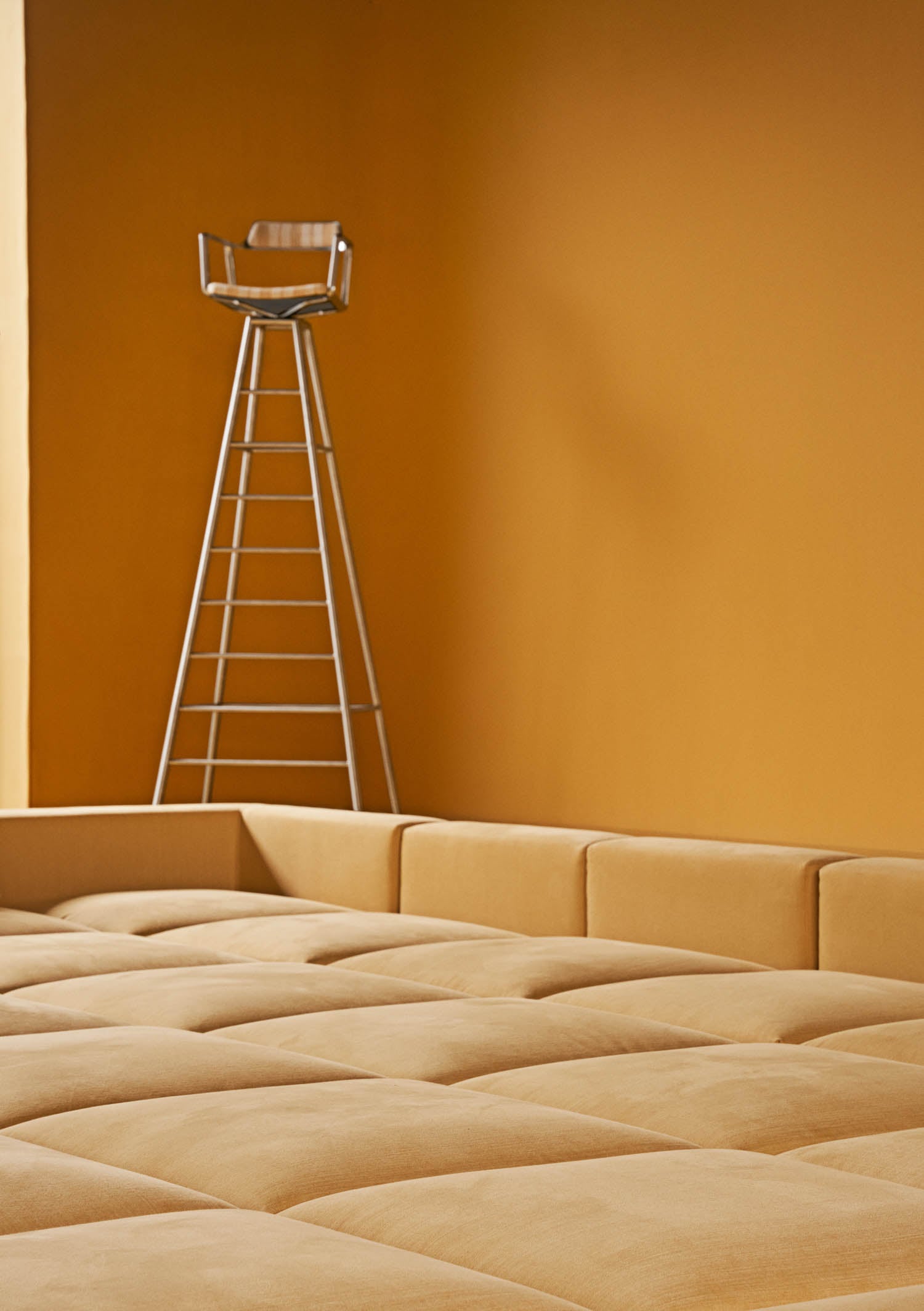 A tall metal bar stool with a wooden seat stands against a mustard yellow wall, next to a large, tufted, mustard yellow upholstered platform.