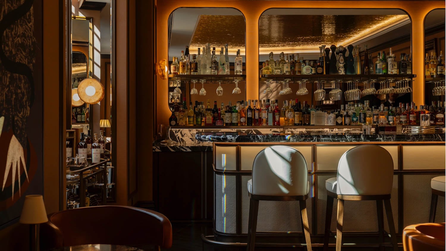 Elegant bar interior with backlit shelves displaying various liquor bottles and glasses, marble countertop, and two white bar stools in the foreground. Warm, ambient lighting throughout.