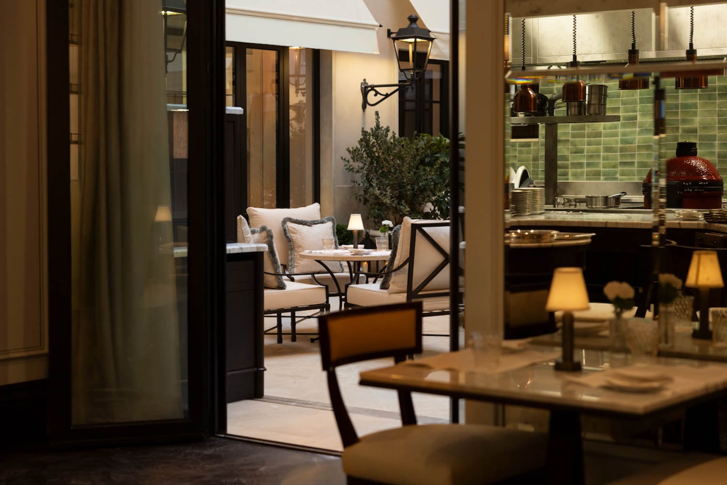 A view from inside a restaurant showing marble tables, cushioned chairs, small lamps, and an open patio area with outdoor seating and greenery.
