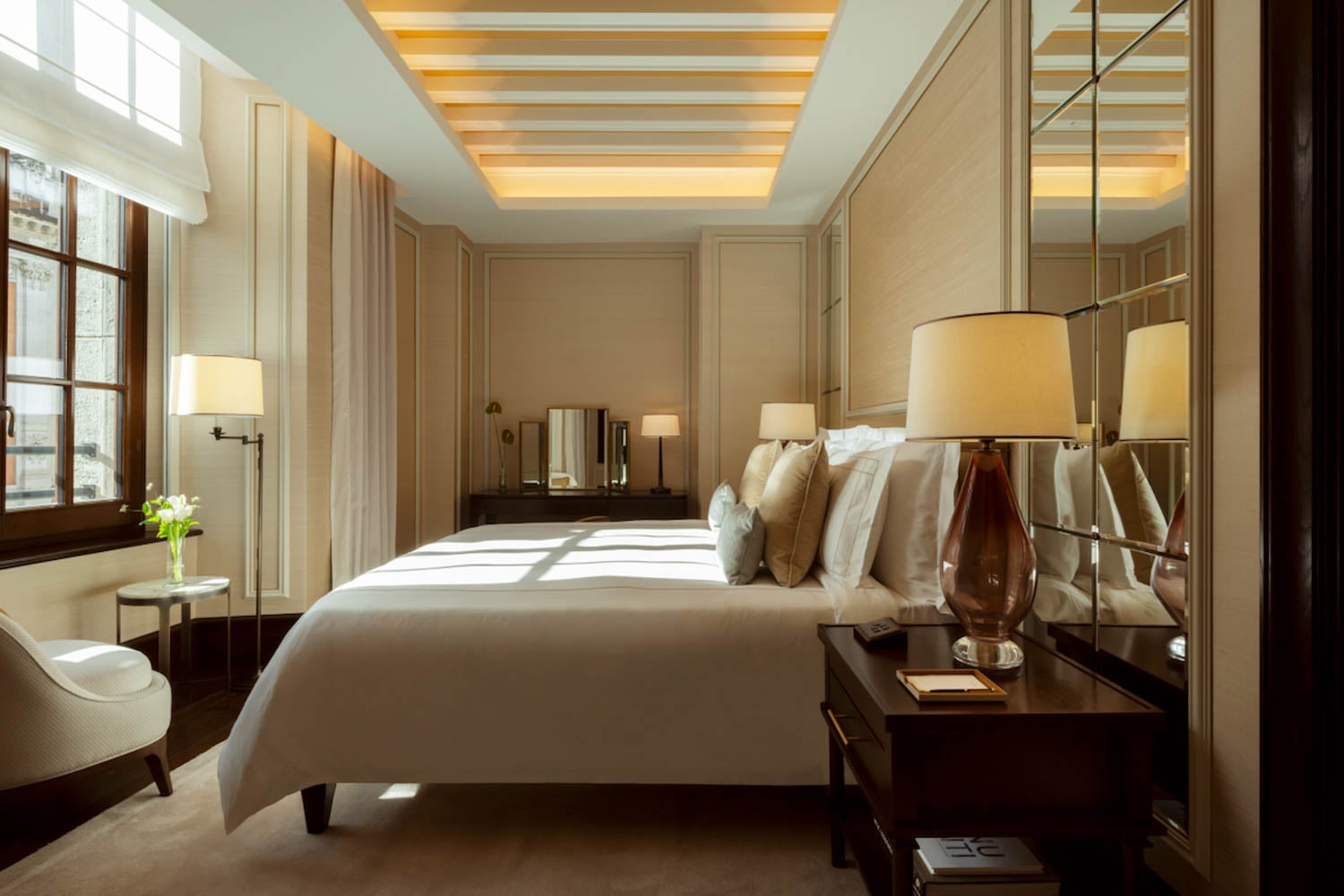 Modern hotel room with a large bed, bedside tables with lamps, cushioned chair, floor lamp, and mirrored walls, illuminated by ceiling and natural light from a window.