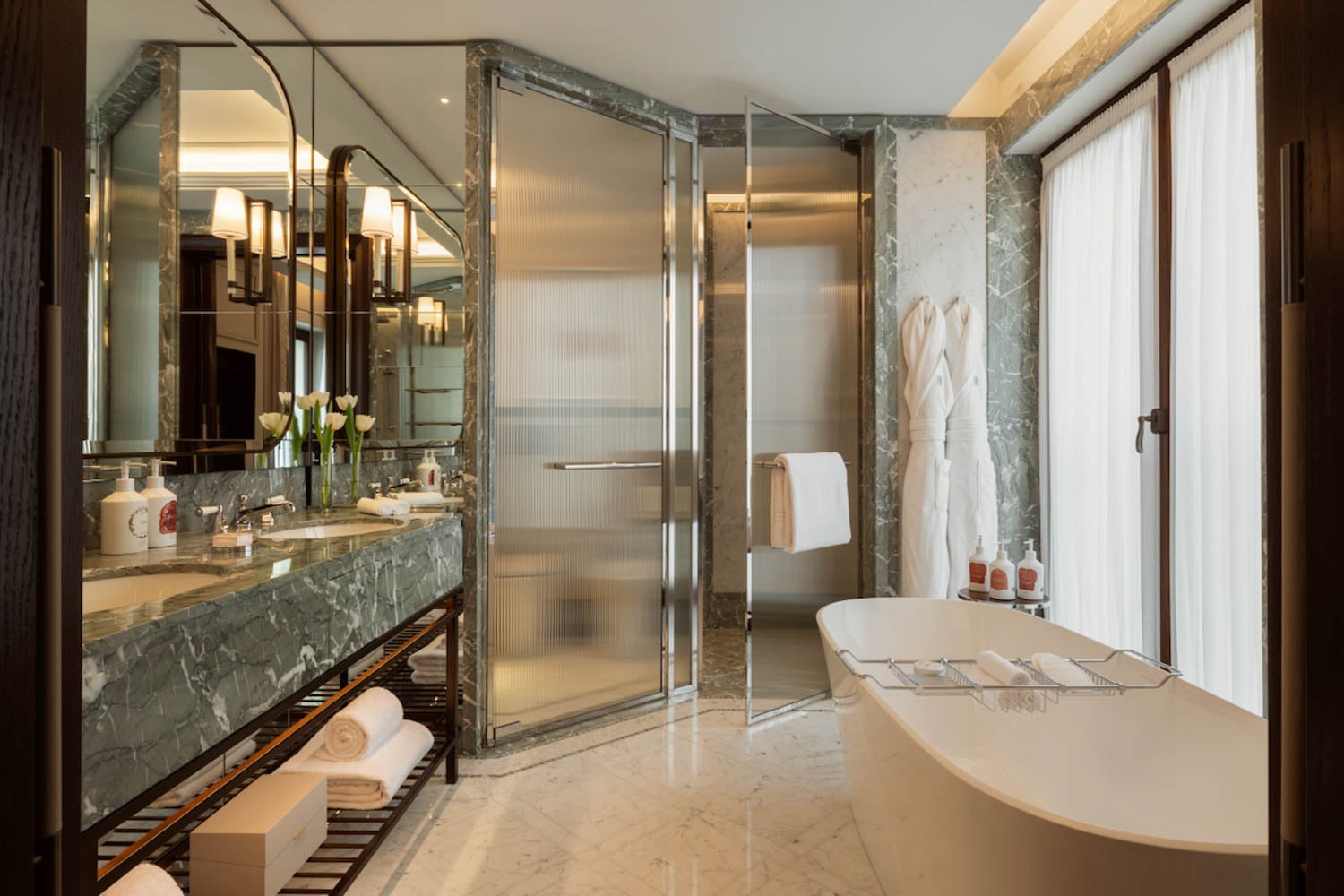 Modern bathroom with marble surfaces, a freestanding bathtub, glass shower, double vanity with mirrors, fresh towels, and two bathrobes hanging near large windows with curtains.