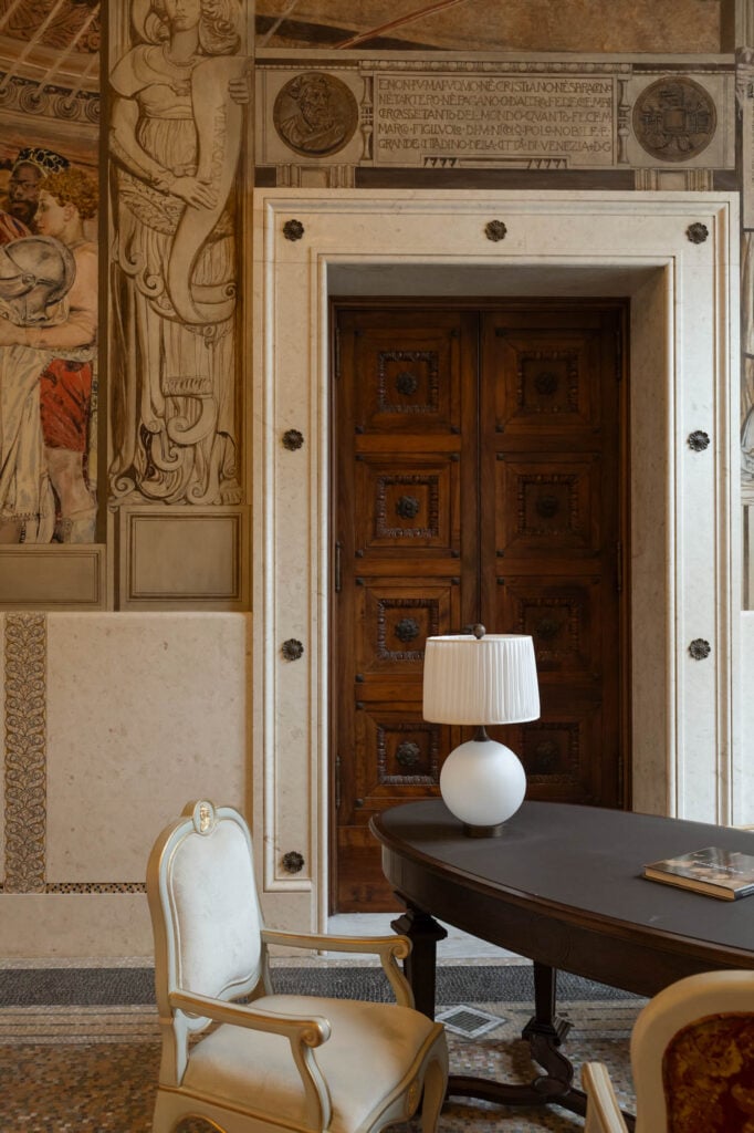 Ornate wooden double doors set in a marble wall, with Renaissance-style mural, a white table lamp, a dark wooden desk, and a cream upholstered chair in the foreground.