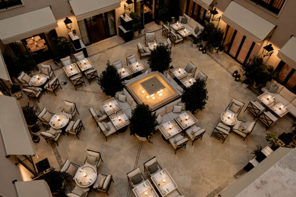 Aerial view of a courtyard restaurant with multiple square tables, cushioned chairs, lit candles, and a central square fire pit surrounded by plants.