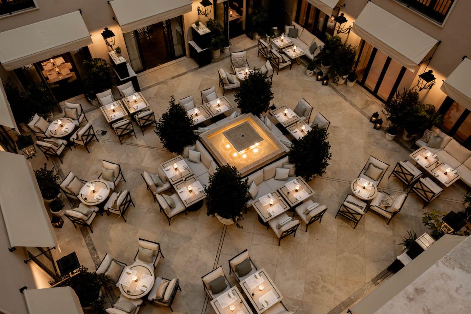Aerial view of a courtyard restaurant with multiple square tables, cushioned chairs, lit candles, and a central square fire pit surrounded by plants.