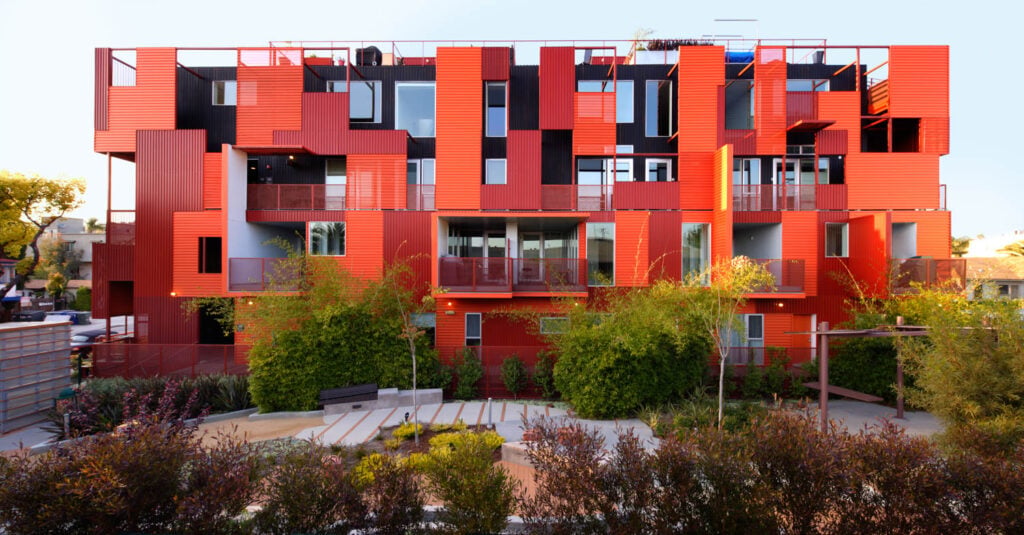 A modern apartment building by Lorcan O'Herlihy with a bold red geometric facade, large windows, and balconies, surrounded by greenery and landscaped outdoor seating areas.