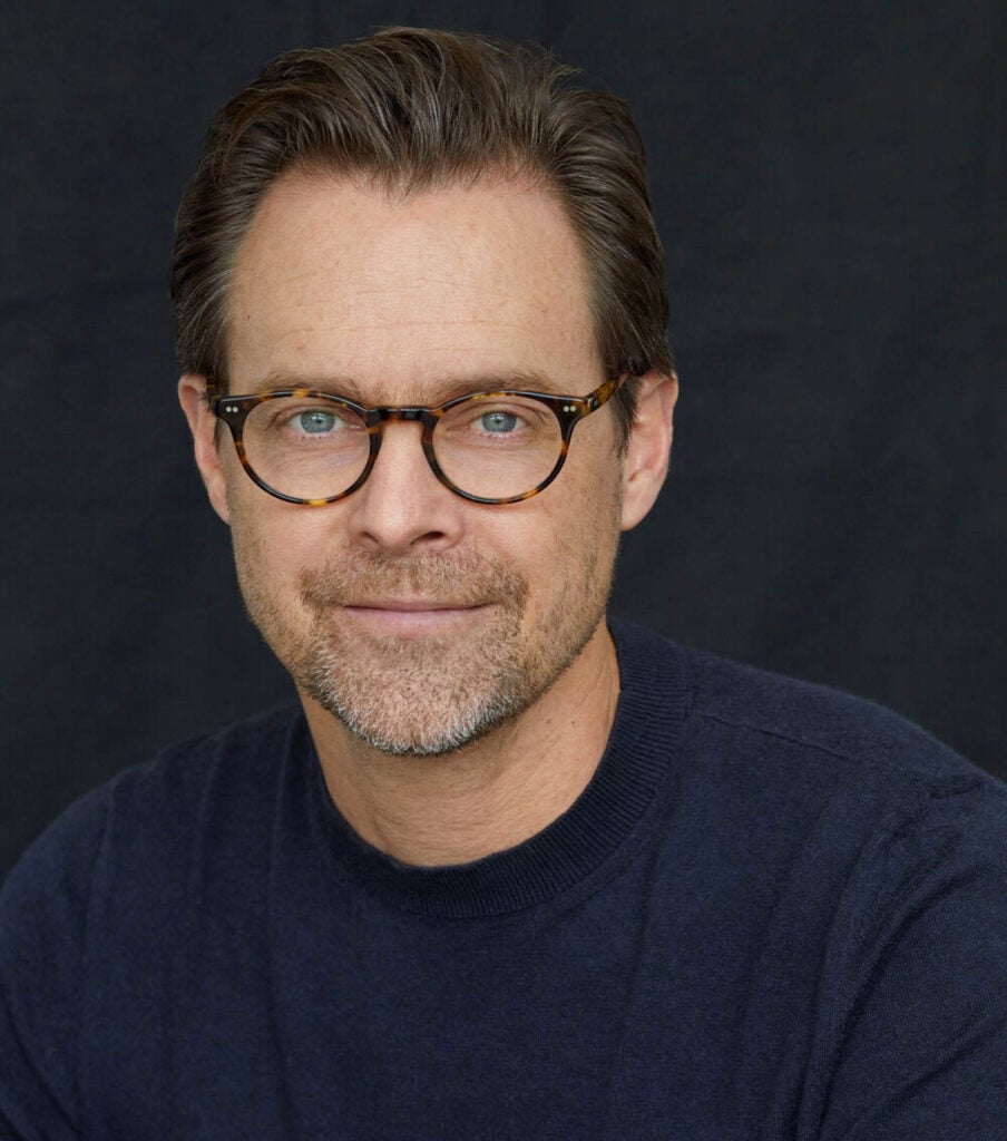 A man with light brown hair, glasses, and a short beard is wearing a navy sweater and looking at the camera, with a plain dark background.