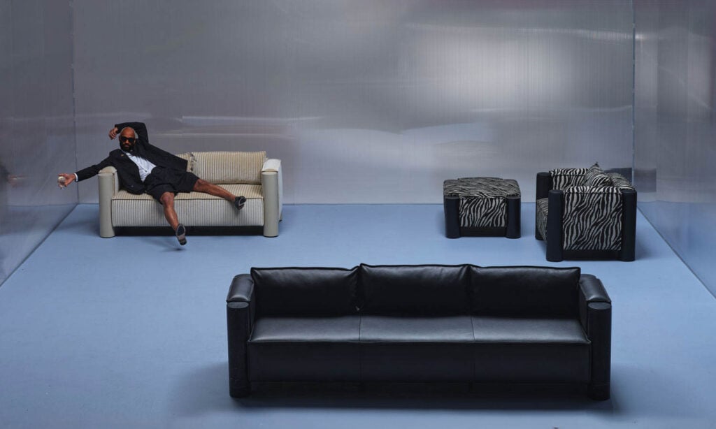 A man in a suit and shorts reclines sideways on a cream sofa in a minimalist room with a black sofa, an armchair, and a zebra-print ottoman.