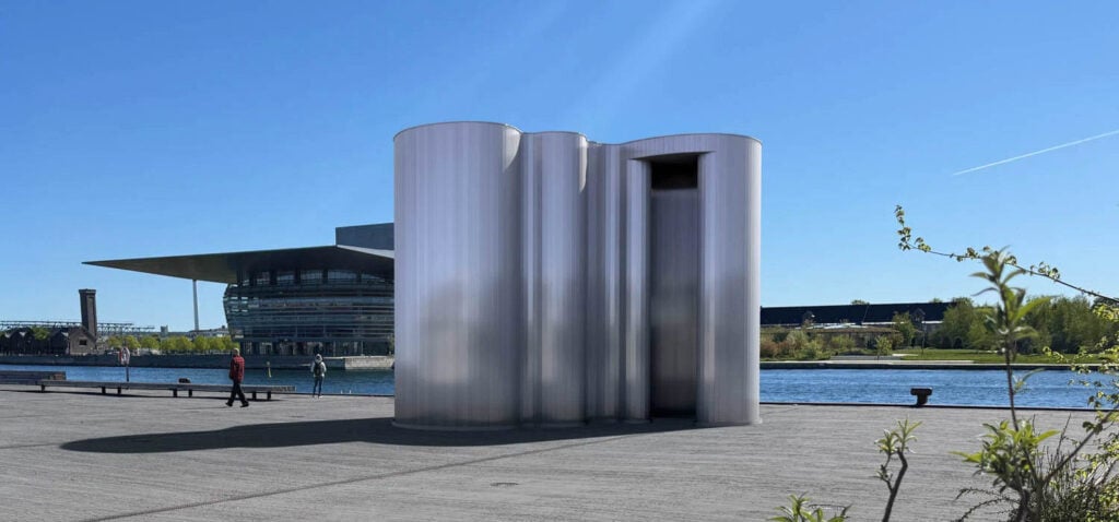 A cylindrical metallic public restroom stands on a paved area near a river, its sleek design reminiscent of 3daysofdesign, with modern buildings and a clear blue sky in the background.