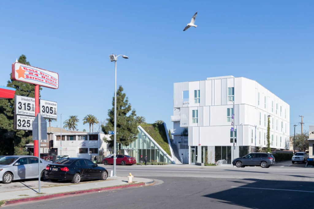 A white modern apartment building, designed by Lorcan O’Herlihy, stands on a sunny street corner next to a gas station, with cars parked nearby and a seagull soaring in the blue sky.