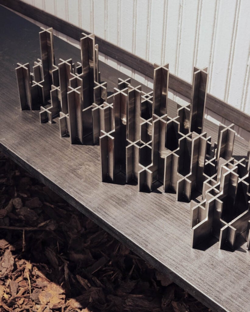 Metal rectangular tubes of varying heights are arranged vertically on a dark surface, resembling a miniature city skyline, with a white wall in the background.