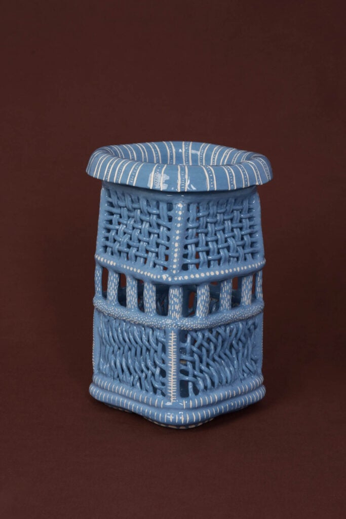 A blue woven stool with a round top, vertical and horizontal slats, and white accent lines, set against a dark brown background.