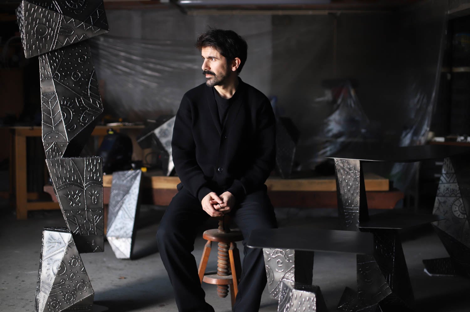 A man in dark clothing sits on a wooden stool in a workshop, surrounded by geometric metal sculptures with engraved patterns.