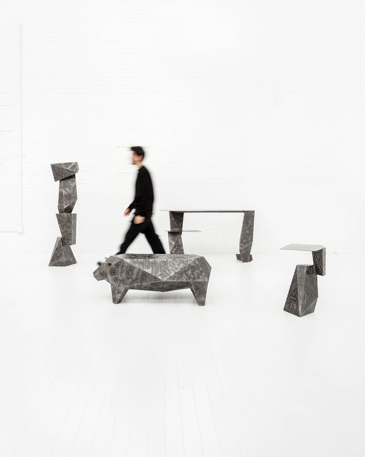 A person in black walks past geometric, sculptural metal furniture pieces in a bright, minimalist white room.
