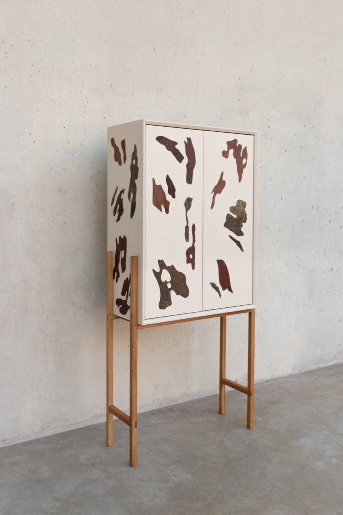 A modern white cabinet with wood legs and abstract brown and rust-colored patterns on its doors and sides, placed against a plain concrete wall.