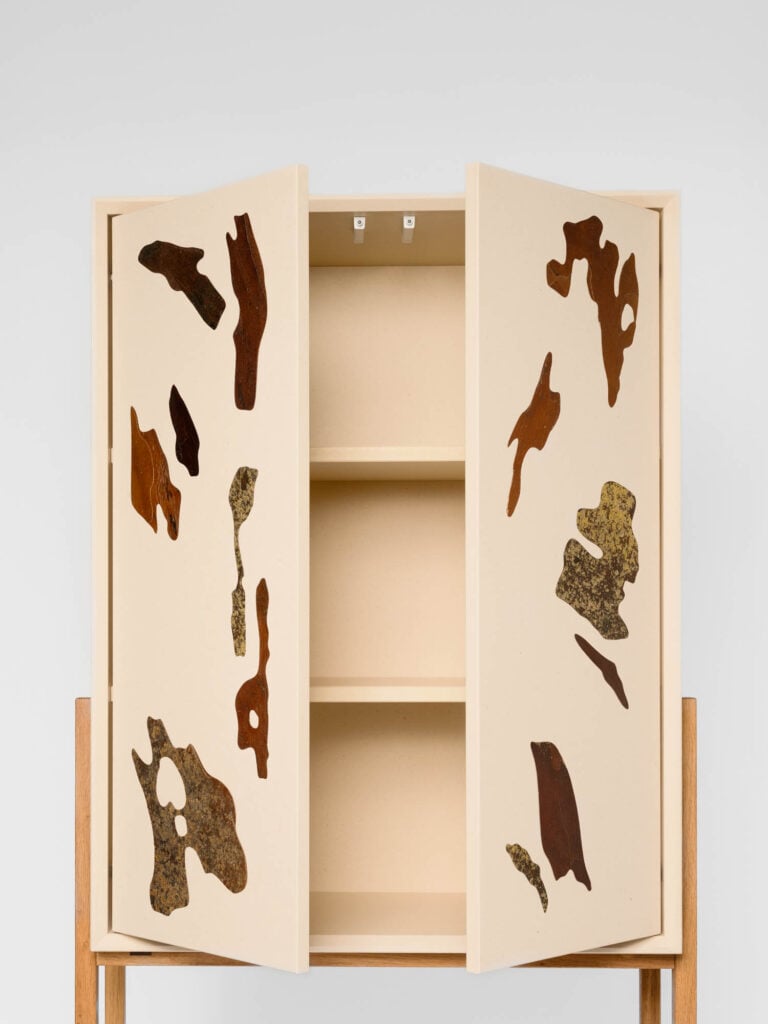 A beige cabinet on wooden legs with abstract brown and green textured shapes on its open doors, revealing three empty shelves inside.