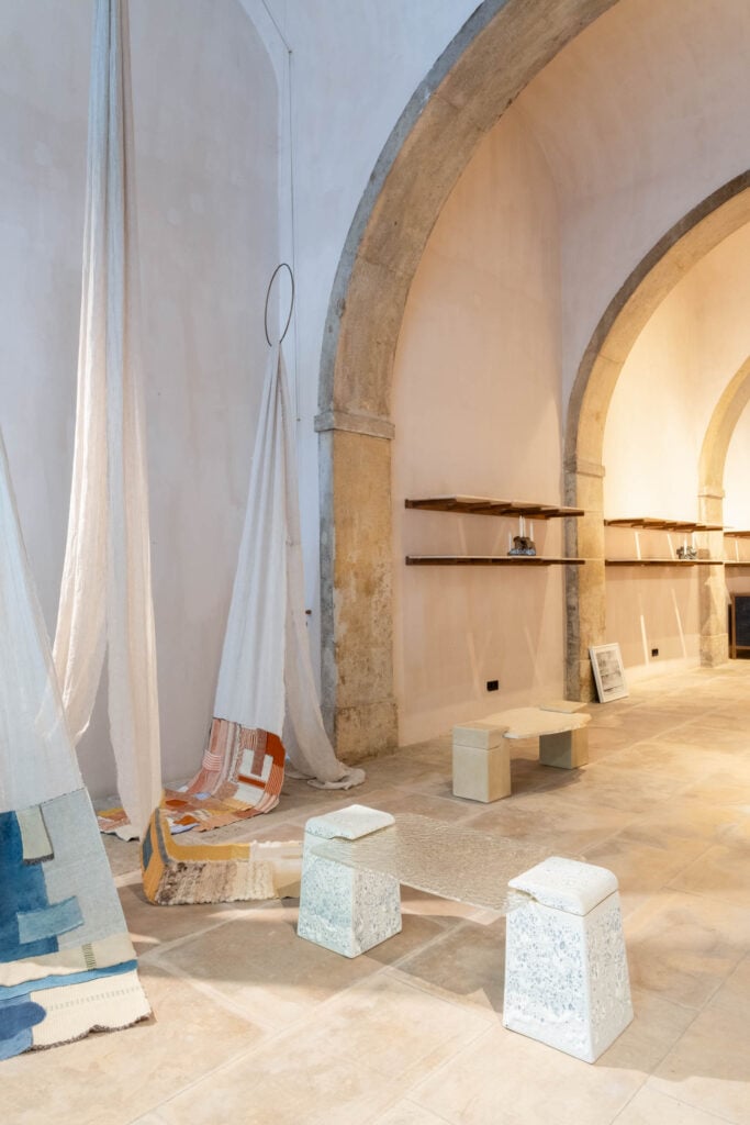 Minimalist room with arched stone walls, two hanging textile installations, three textured stools, a small table, and floating wooden shelves with decorative items.