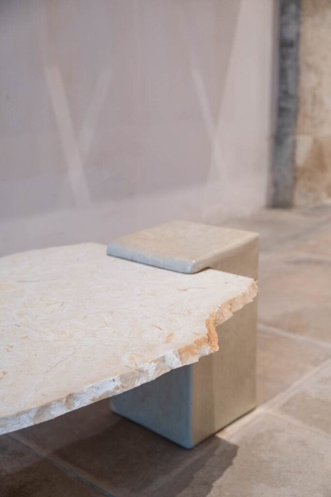 A close-up of a modern, minimalist stone bench with a rough-edged seat and geometric supports set on a tiled floor.