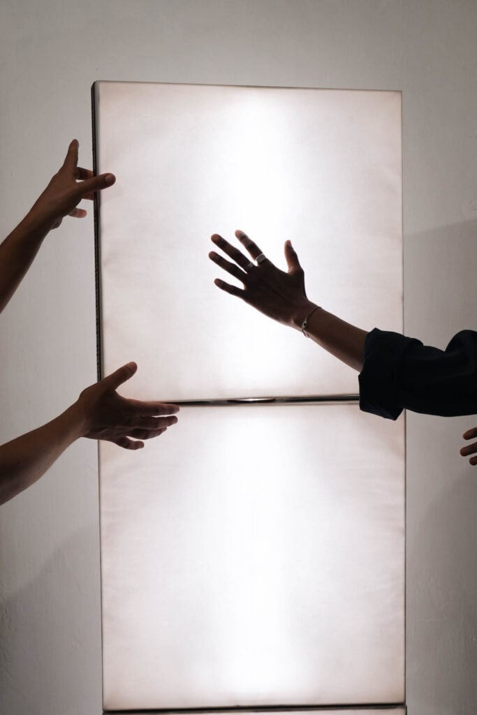 Two pairs of hands reach toward a brightly lit, frosted glass panel positioned vertically against a plain white background.
