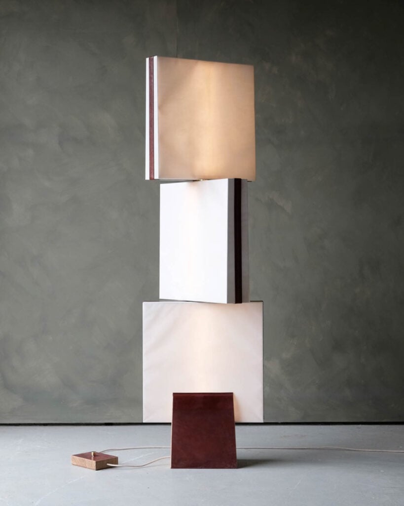 A modern floor lamp with three illuminated square panels stacked vertically on a rectangular brown base, set against a plain grey wall.